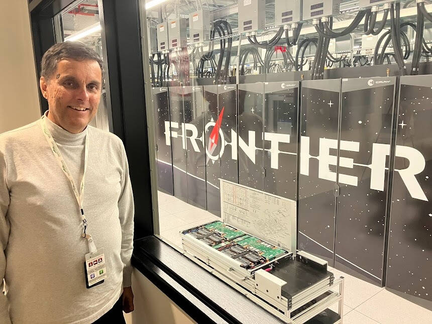 A smiling man stands beside a large window or glass wall, looking into the Oak Ridge National Laboratory data center where the “Frontier” supercomputer is installed behind panels labeled “FRONTIER.”